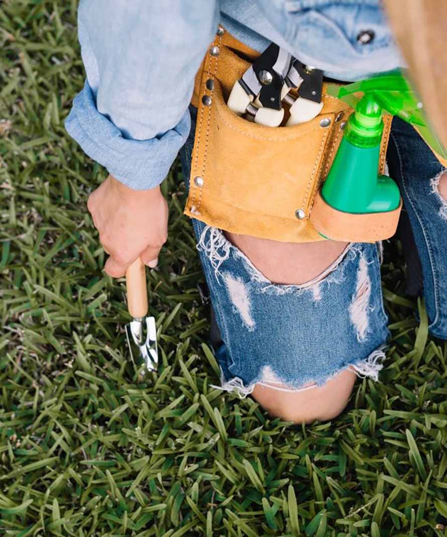 Gardener kneeling in grass with a tool belt holding pliers and a spray bottle at the hip; hand trowel in use.
