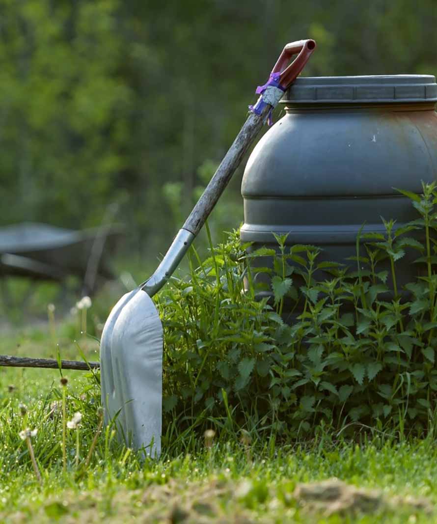 Shovel propped against a large gray rain barrel in a garden with tall weeds and green grass.