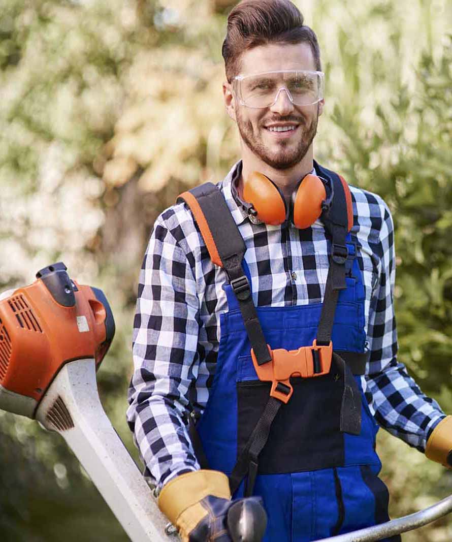 Gardener wearing safety goggles, orange ear protection, and blue overalls, holding a leaf blower outdoors.
