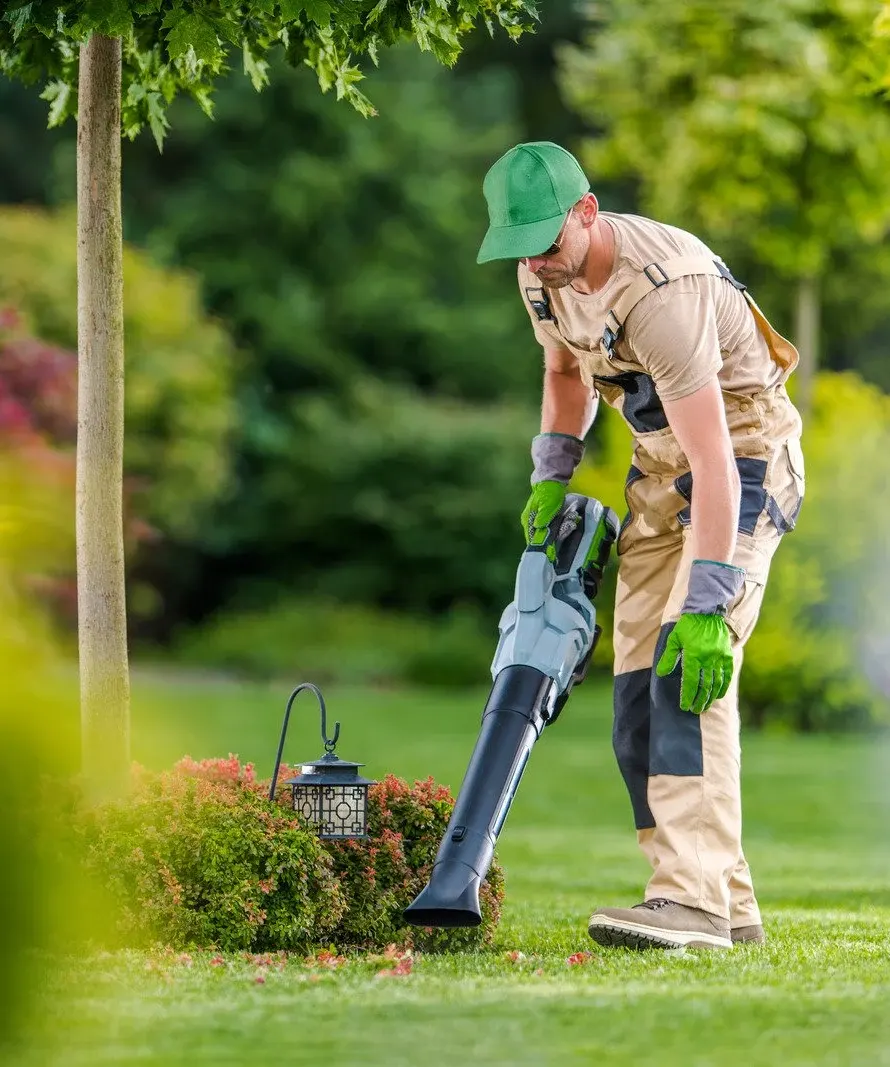 Gardener wearing a green cap and beige work overalls uses a leaf blower to clear debris from a small garden bed.
